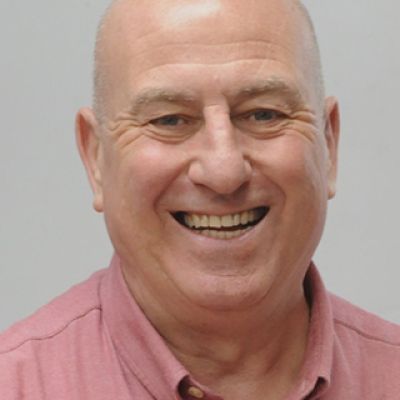 Head and shoulders shot of a smiling Richard Jones wearing a pink shirt stood in front of a grey wall looking into the camera